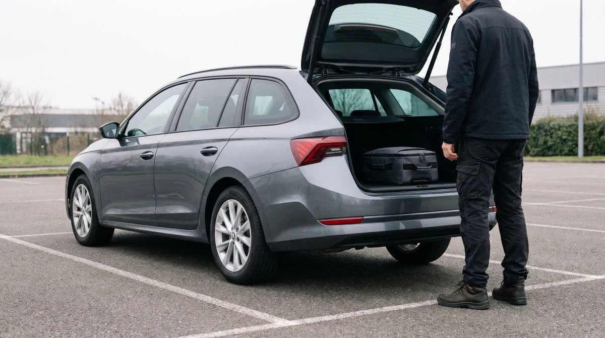 Un homme se tient devant le coffre ouvert d’un break gris chargé d’une valise, sur un parking.