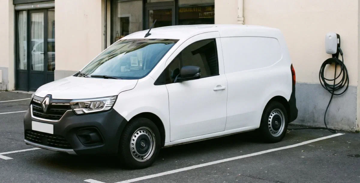Un utilitaire blanc est garé sur une place de parking, à côté d’une borne de recharge fixée au mur.
