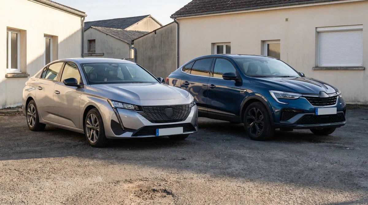 Une berline grise et un SUV coupé bleu garés côte à côte dans la cour d’un bâtiment d’entreprise.
